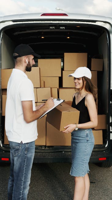 Two men wearing black face masks are engaged in a conversation beside a white moving van with its rear doors open, revealing packed cardboard boxes, some labeled with packing tape, and miscellaneous i